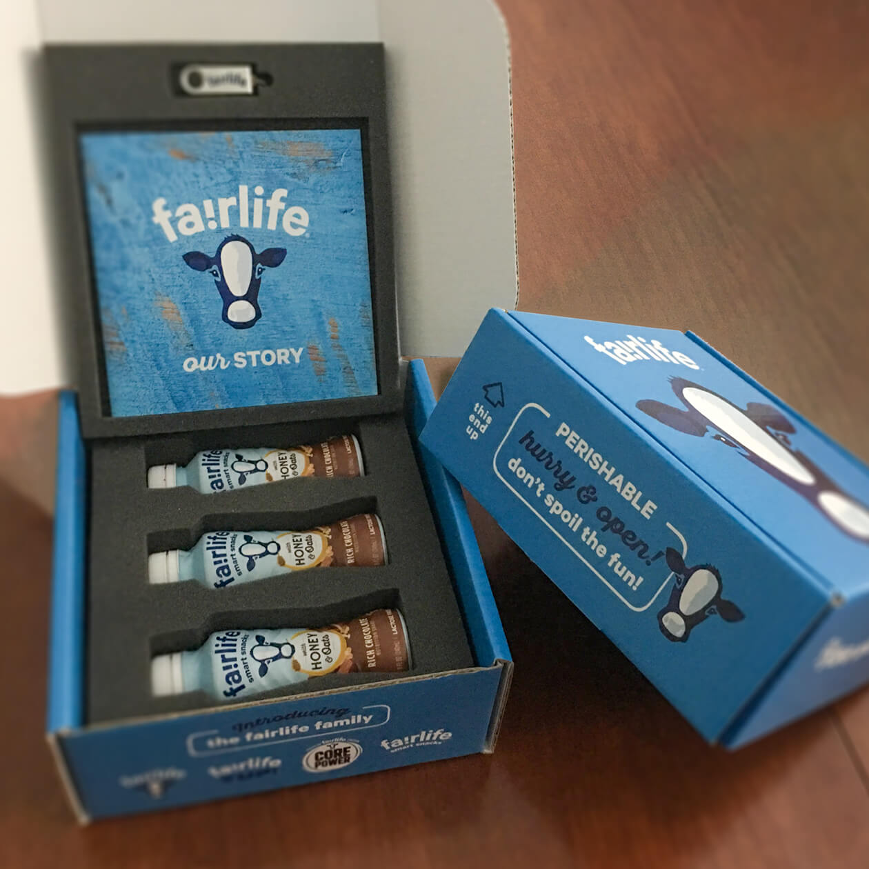 opened fairlife blue Press Kit with cow head on brown wood background, kit contains brochure, custom USB loaded with fact sheets and fairlife smart snacks product line